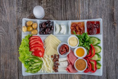 Gri ahşap masada geleneksel Türk kahvaltı tabağı, üst görünüm: pogaca hamur işleri, sebzeler, peynir, zeytin ve helal hindi salamı, seçici odak noktası; metin için kopyalama alanı.