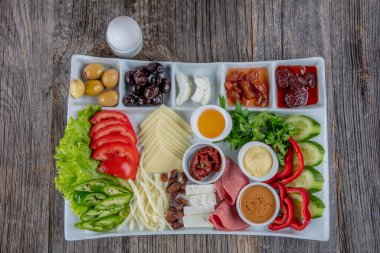 Gri ahşap masada geleneksel Türk kahvaltı tabağı, üst görünüm: pogaca hamur işleri, sebzeler, peynir, zeytin ve helal hindi salamı, seçici odak noktası; metin için kopyalama alanı.