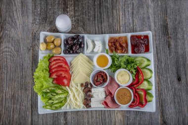 Gri ahşap masada geleneksel Türk kahvaltı tabağı, üst görünüm: pogaca hamur işleri, sebzeler, peynir, zeytin ve helal hindi salamı, seçici odak noktası; metin için kopyalama alanı.