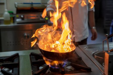 Kızartma Tavasında Ateşle Pişirilen Şef. Ticari bir mutfakta profesyonel bir aşçı alevli yemek pişirme stili. Aşçı, ticari mutfakta alevli tavada yemek pişiriyor..