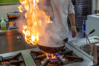 Kızartma Tavasında Ateşle Pişirilen Şef. Ticari bir mutfakta profesyonel bir aşçı alevli yemek pişirme stili. Aşçı, ticari mutfakta alevli tavada yemek pişiriyor..