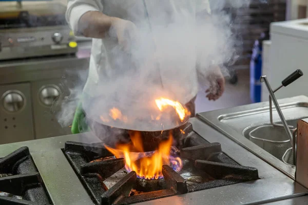 Chef Cooking With Fire In Frying Pan. Professional chef in a commercial kitchen cooking flambe style. Chef frying food in flaming pan on gas hob in commercial kitchen.