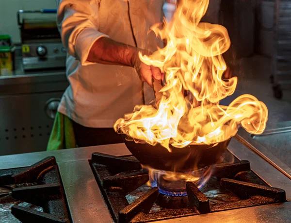 Chef Cooking With Fire In Frying Pan. Professional chef in a commercial kitchen cooking flambe style. Chef frying food in flaming pan on gas hob in commercial kitchen.