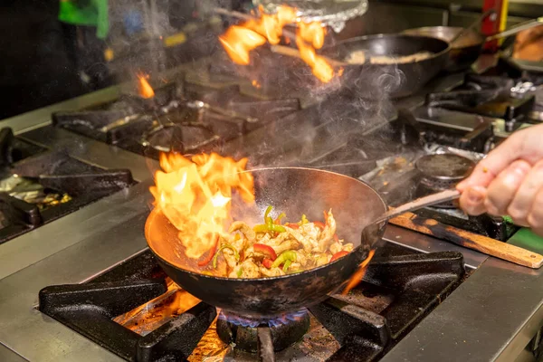 Chef Cooking With Fire In Frying Pan. Professional chef in a commercial kitchen cooking flambe style. Chef frying food in flaming pan on gas hob in commercial kitchen.