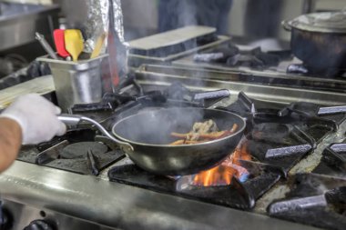 Kızartma Tavasında Ateşle Pişirilen Şef. Ticari bir mutfakta profesyonel bir aşçı alevli yemek pişirme stili. Aşçı, ticari mutfakta alevli tavada yemek pişiriyor..