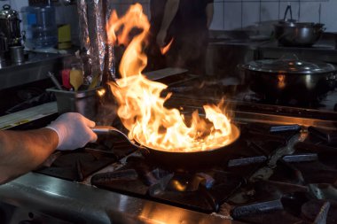 Kızartma Tavasında Ateşle Pişirilen Şef. Ticari bir mutfakta profesyonel bir aşçı alevli yemek pişirme stili. Aşçı, ticari mutfakta alevli tavada yemek pişiriyor..