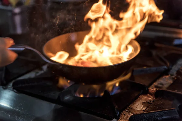 Chef Cooking With Fire In Frying Pan. Professional chef in a commercial kitchen cooking flambe style. Chef frying food in flaming pan on gas hob in commercial kitchen.