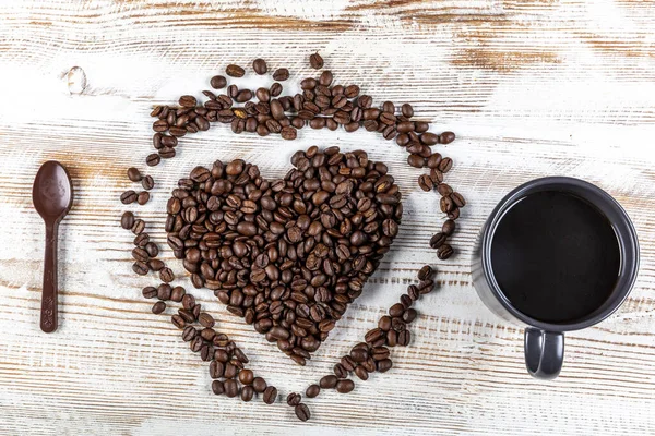 Cup of coffee, heart shape made from coffee beans and chocolate spoon on wooden background. Symbol of Love to coffee.