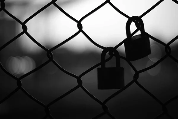 Silhouette of two pad locks on chain link fence against bright background.