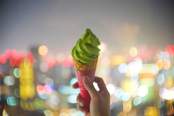 Soft focus hand holding sweet yummy matcha green tea ice-cream and crispy crunchy cone with defocused of colorful bokeh street city light night shiny glow decoration blurred festival party background.