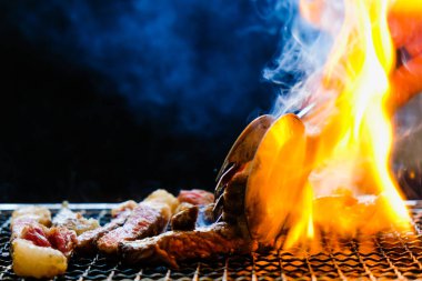 Gümüşle servis edilen mutfak maşası, çiğ biftek, dilimlenmiş bbq ızgara kömür sobası, duman ve siyah arka planda ateş..