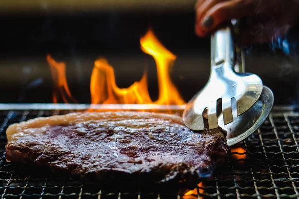 Gümüşle servis edilen mutfak maşası, çiğ biftekli bbq ızgarası, kömür ocağı dumanı ve siyah arka planda ateş alevi..