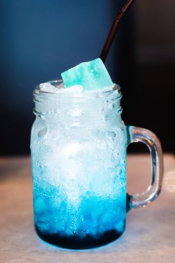 Yakın plan mason kavanozu. Soğuk, mavi lagün kokteyli. Masada buz ve bulanık koyu arkaplan..