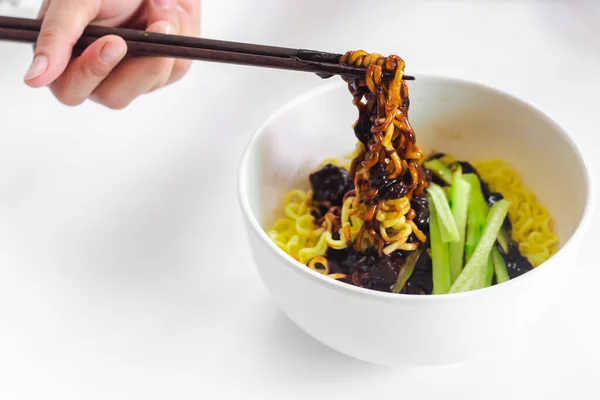 韓国の黒豆麺を持つ箸を使う手 白鉢の中に黒豆と野菜を入れたJajangmyeon麺と白背景の箸 — ストック写真