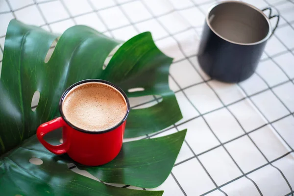a red cup of coffee on green leaf and black cup on white grid fabric background.