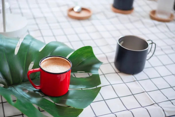 a red cup of coffee on green leaf and black cup on white grid fabric background.