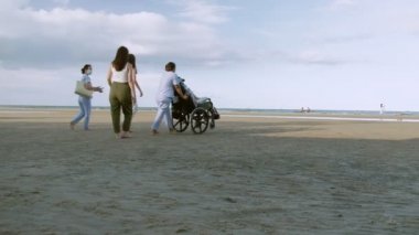 Tekerlekli sandalyedeki genç adam, hemşire ve ailesiyle birlikte deniz kenarındaki kumsalda tatilin tadını çıkarıyor..