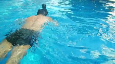 Rahatlayan genç adam, yaz tatili tatilinde gün ışığı ve su yüzeyinin yansımasıyla yüzme havuzunda temiz suyun tadını çıkarıyor..