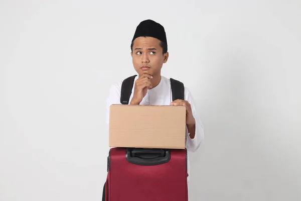 Portrait of confused Asian muslim man holding suitcase and cardboard box and thinking with hand on chin. Going home for Eid Mubarak. Isolated image on white background