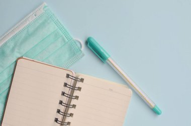 Mavi arka planda kalem ve tıbbi maskeli bir defter. Üst Manzara. Boşluğu kopyala
