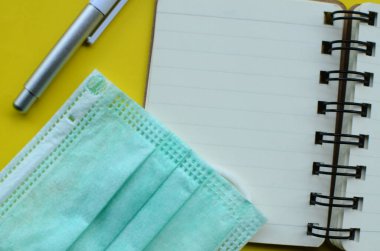 Yüzü maskeli boş bir defter ve sarı arka planda bir kalem..