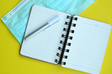 Yüzü maskeli boş bir defter ve sarı arka planda bir kalem..