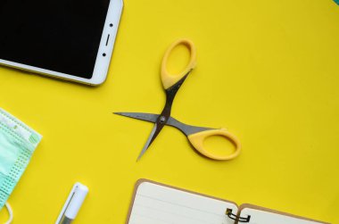 Yüzünde tıbbi maskeler, cep telefonları, defterler, makas, sarı arka planda kalemler olan düz bir kompozisyon. Kopyalama alanı olan üst görünüm.