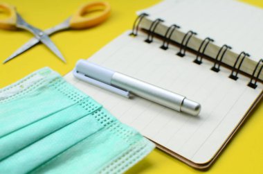 Yüzü maskeli boş bir defter ve sarı arka planda bir kalem..