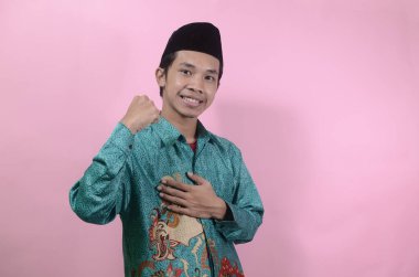 Genç adam zafer kutlamalarında, kazanan bir jest gösteriyor. Pembe arka planda izole edilmiş batik gömlek ve kafatası kapağı giyen Asyalı genç bir adamın portresi.