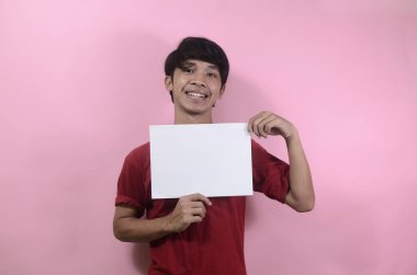 Genç adam yazı ve metin için boş bir poster tutuyor. Kırmızı tişört giyen Asyalı adamlar pembe arka planda izole edilmiş.