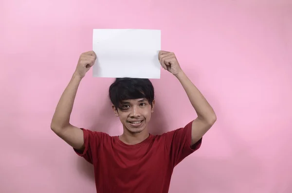 这个年轻人拿着一张空白的招贴画 上面写着和文字 亚洲男人穿着红色T恤 背景是粉红色的 — 图库照片