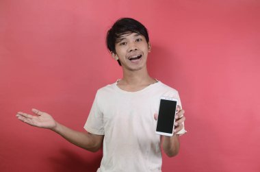 Mutlu gülümseyen Asyalı genç adam cep telefonunu gösteriyor. Kırmızı arka planda beyaz tişörtler giyen Asyalı adamlar.