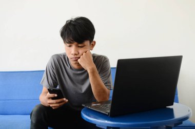 Asyalı gençler dizüstü bilgisayarın önündeki koltukta cep telefonuyla oturuyorlar.