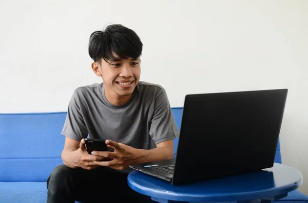 Asian youth sitting on the sofa in front of the laptop using a cell phone