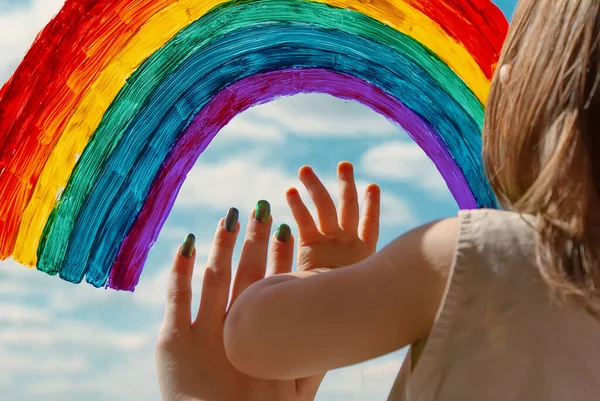 children\'s hands on the window pane next to a painted colored rainbow. symbol of happiness and love