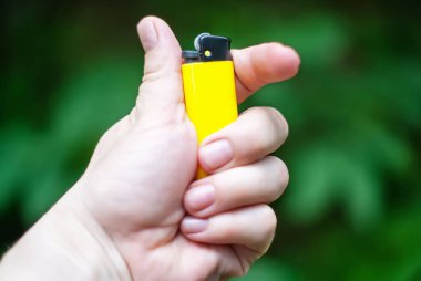 Sokakta bir kadının eli, arka planda yeşil yapraklarla kaplı bir çakmağı tutuyor. Bir kadının elinde sarı bir çakmak.