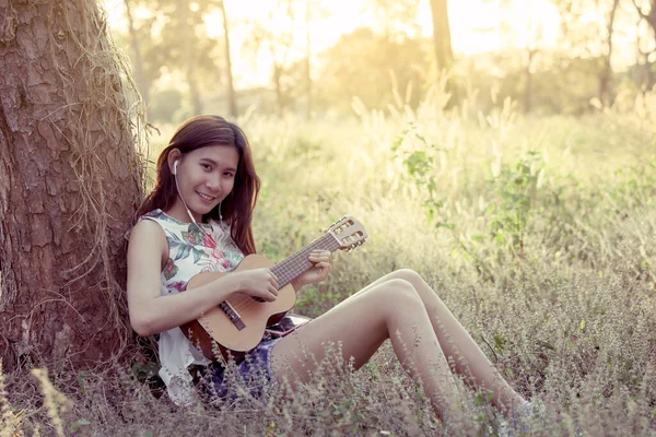 Young asian woman put on earphones listening music and playing acoustic guitalele in the park in vintage color tone