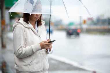 Asyalı kadın akıllı telefon kullanıyor, sosyal medya ağını kontrol ediyor ve yağmurlu bir günde şehir caddesinde taksi beklerken şemsiye tutuyor.