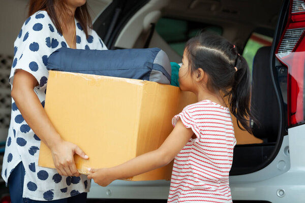 Happy asian child girl and mother helping to carry a cardboard box with stuffs moving into the car together to relocation on moving day. Home renovation and relocation concept.