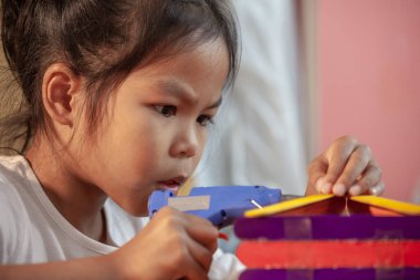 Asyalı çocuk, sıcak eritilmiş elektrikli yapıştırıcı tabancasıyla renkli dondurma çubukları yapıştırıyor. Çocuklar bir el işi projesinde ev yapmak için eğleniyorlar..