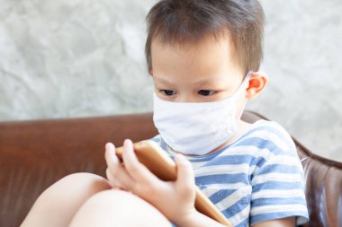 Korunma maskesi takan Asyalı çocuk, Coronavirus Covid-19 ve hava kirliliği pm2.5 nedeniyle ev karantinasındaki akıllı telefondan oynuyor..