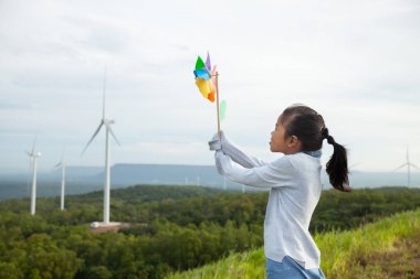 Rüzgâr türbini tarlasında elinde rüzgar oyuncağıyla oynayan Asyalı bir çocuk..