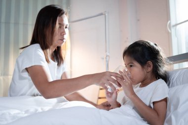 Asyalı anne, hasta kıza yatak odasında ilaç içtikten sonra bir bardak su veriyor. Hasta çocuk ve sağlık hizmeti kavramı.