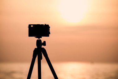 Tripodun üzerinde eski bir kamera silueti güneşin doğuşunda ya da batışında suya yansıyan güneşli sakin bir denizi çekiyor.