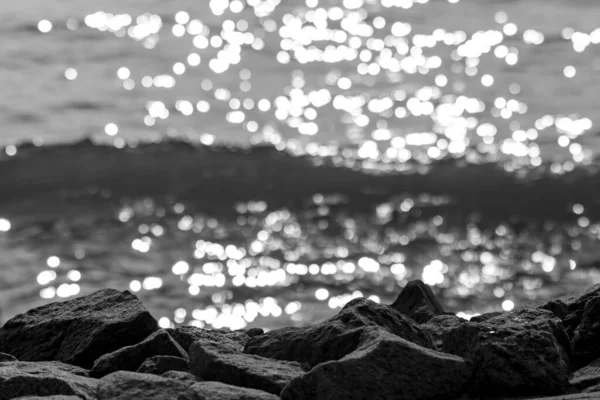 Güneş batarken deniz kenarındaki kayalar su dalgalarını, siyah ve beyaz fotoğrafçılığı yansıtır.