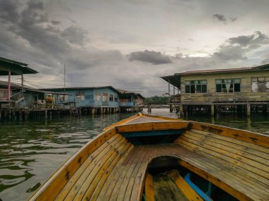 Hareket halindeki bir tekneden Bandar Serisi Begawan Brunei Darussalam 'daki su köyündeki ahşap evlerin görüntüsü. Arkaplanda bulutlu bir gökyüzü, ahşap evlerin odağı.