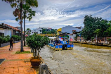 Malacca City, Malezya - 19 Aralık 2019: Yürüme yolu, nehir kenarındaki yenilenmiş binalar, mavi gökyüzü görüş mesafesi ve hareket halindeki tekne bulanıklığı ile Malacca nehri. Önplanda seçmeli odak.