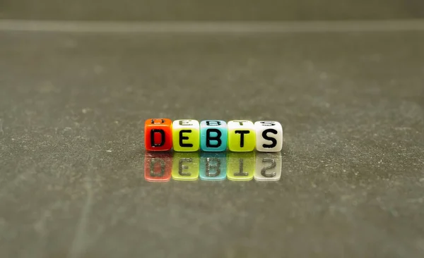 Conceptual of debts, an item in financial report or audited accounts spelled on colorful alphabet beads. Isolated over reflective dark background. Selective focus on beads; other in gradient blur.
