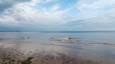 Deniz manzarasında, sakin bir deniz, küçük dalgalar ve kumlu sahil kenarında nadir küçük dalgalar, gökyüzünde gri bulutlar mavi gökyüzünü kaplıyor, ve her şey akşamın altın rengine dönüşmeye başlıyor..