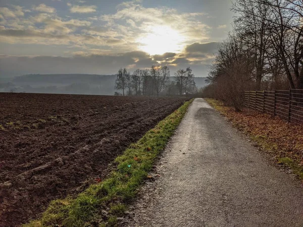 Walking path along plowed field in the afternoon sun in rural Germany, with the sun glowing brilliantly behind clouds and mist in the distance